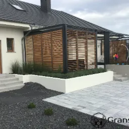 Nowoczesna pergola z drewnianymi lamelami, przylegająca do domu, z widocznym tarasem wyłożonym szarą kostką brukową i ozdobnym żwirem.