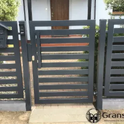 Nowoczesna, grafitowa furtka z poziomymi panelami, zintegrowana ze skrzynką na listy i domofonem, na tle domu z brązowymi drzwiami.