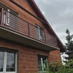 Nowoczesna balustrada balkonowa z motywem labiryntu, wykonana z ciemnobrązowej stali, zamontowana na balkonie w domu z czerwonej cegły, widok z dołu w pochmurny dzień.