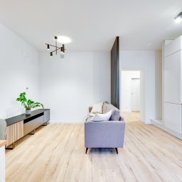 PROFIT Katarzyna Ratajczyk - Jasne, przestronne wnętrze: salon z ceglaną ścianą, szarą sofą, drewnianą podłogą i aneksem kuchennym z białymi szafkami.