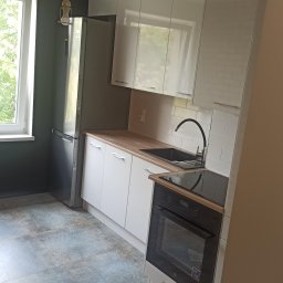 PROFIT Katarzyna Ratajczyk - Nowoczesna kuchnia z białymi, błyszczącymi szafkami, drewnianym blatem i płytkami imitującymi cegłę na ścianie, z widoczną niedokończoną instalacją oświetleniową w postaci wiszącej żarówki.