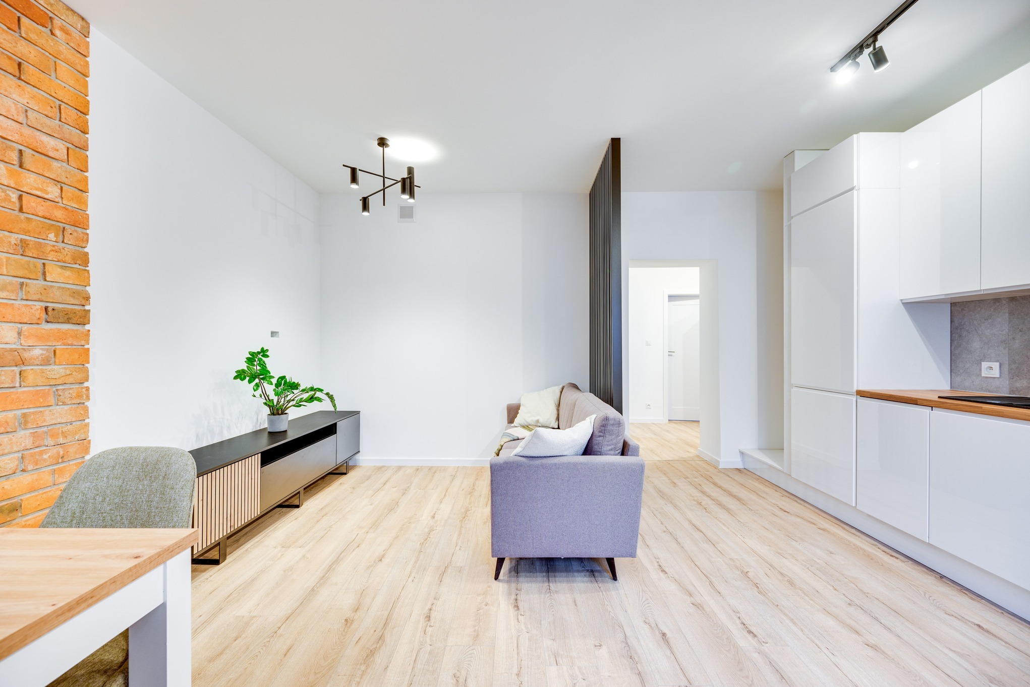 Jasne, przestronne wnętrze: salon z ceglaną ścianą, szarą sofą, drewnianą podłogą i aneksem kuchennym z białymi szafkami.