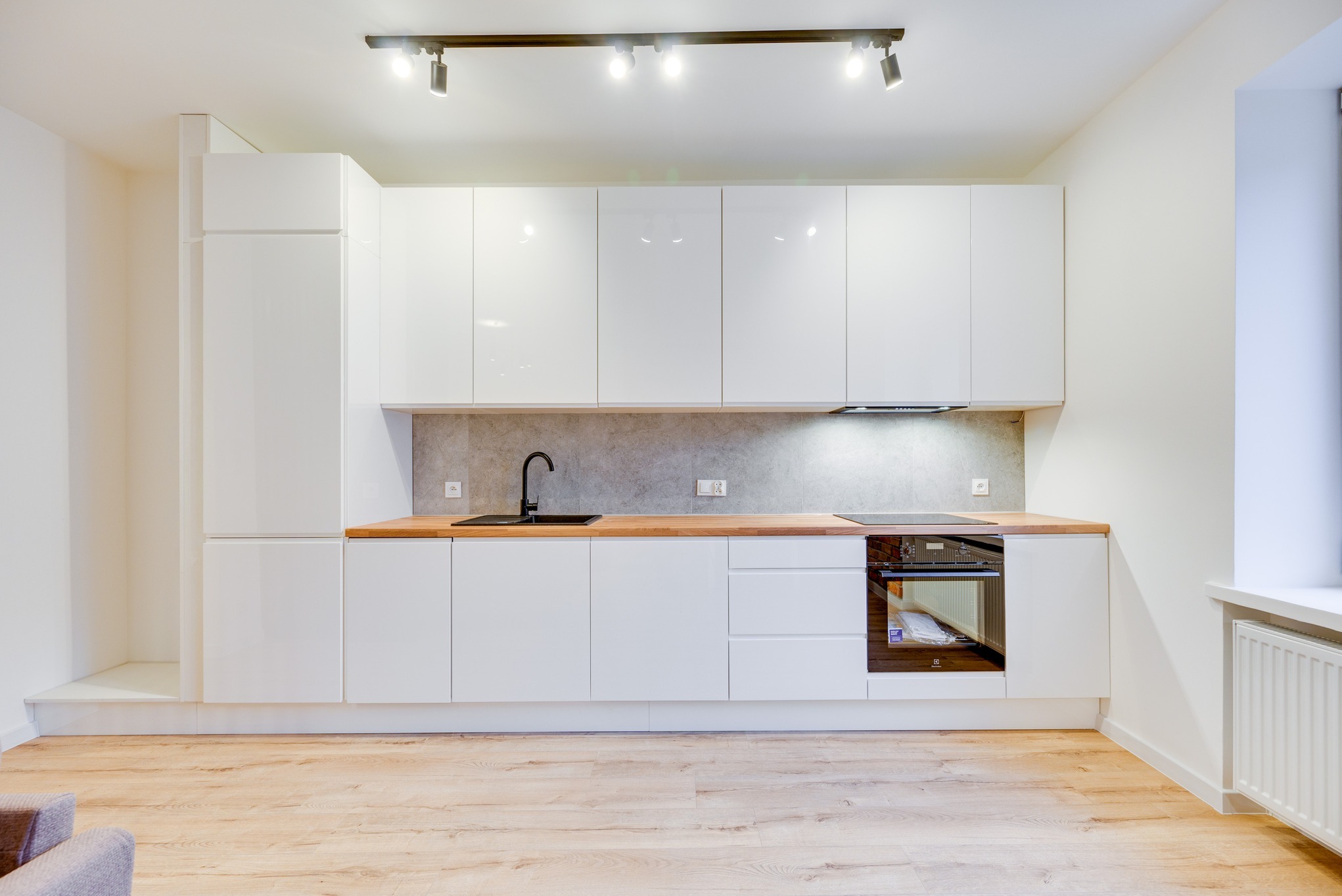 Nowoczesna, minimalistyczna kuchnia z białymi, lakierowanymi szafkami, drewnianym blatem, czarną armaturą i szarym panelem ściennym, oświetlona szynoprzewodem z reflektorami