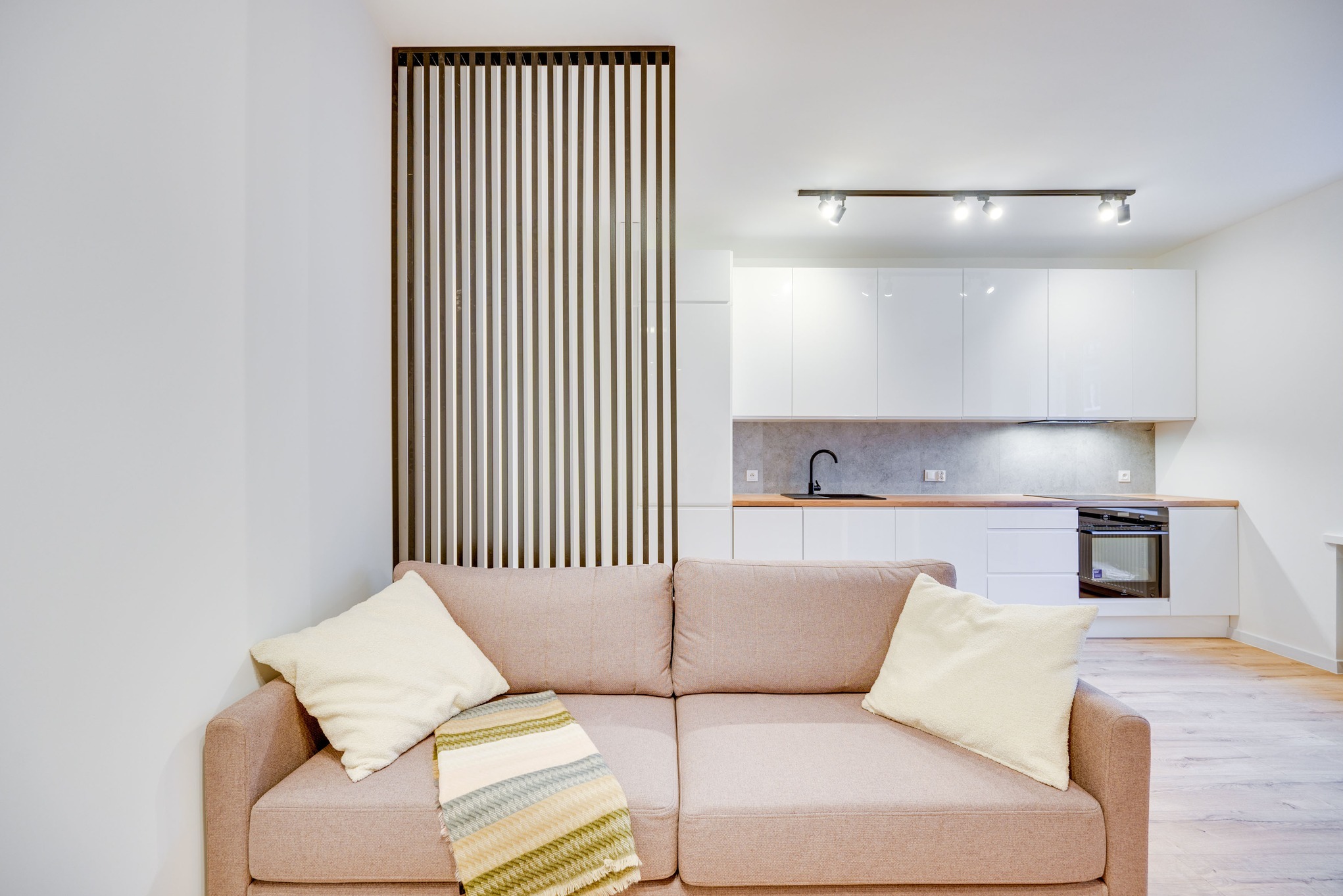 Jasny salon z beżową sofą, kremowymi poduszkami i zielono-białym pledem na tle minimalistycznej, białej kuchni z drewnianym blatem i czarną armaturą, oddzielonej ażurową ścianką z drewnianych listew.