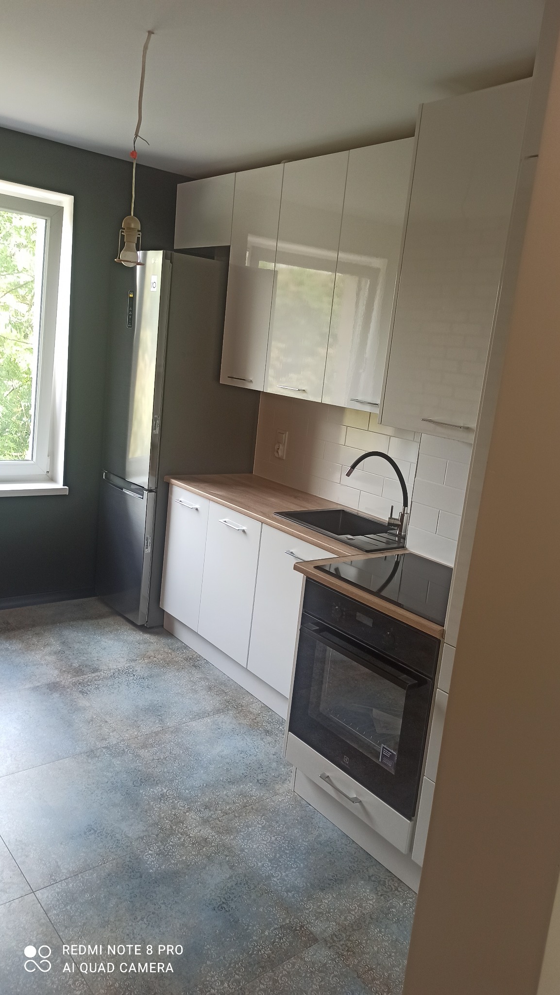 Nowoczesna kuchnia z białymi, błyszczącymi szafkami, drewnianym blatem i płytkami imitującymi cegłę na ścianie, z widoczną niedokończoną instalacją oświetleniową w postaci wiszącej żarówki.