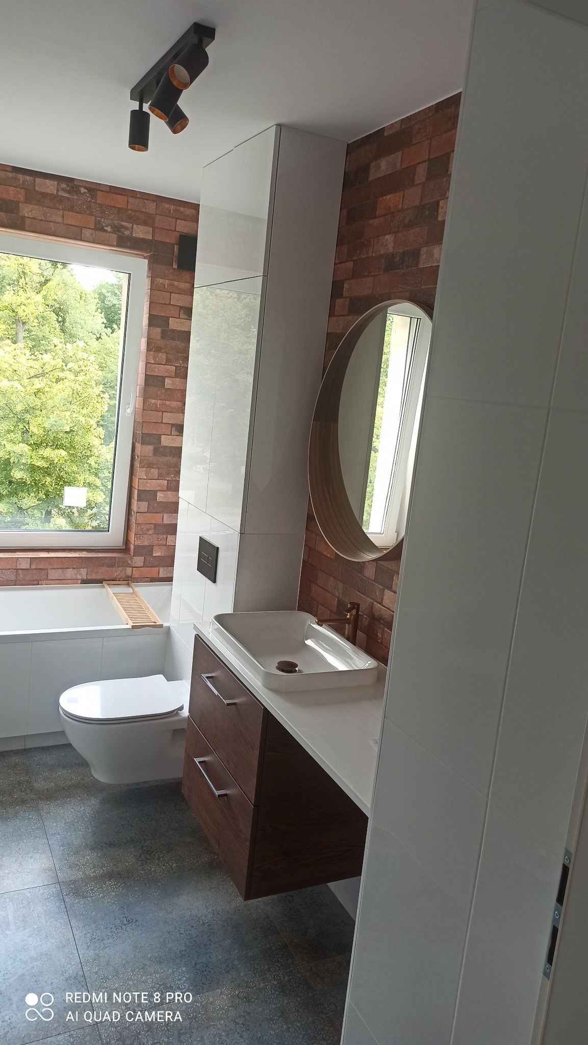 Nowoczesna łazienka z ceglaną ścianą, okrągłym lustrem w złotej ramie, białą umywalką nablatową na drewnianej szafce z szufladami, toaletą i wanną przy oknie z widokiem na zieleń, oświetlona...
