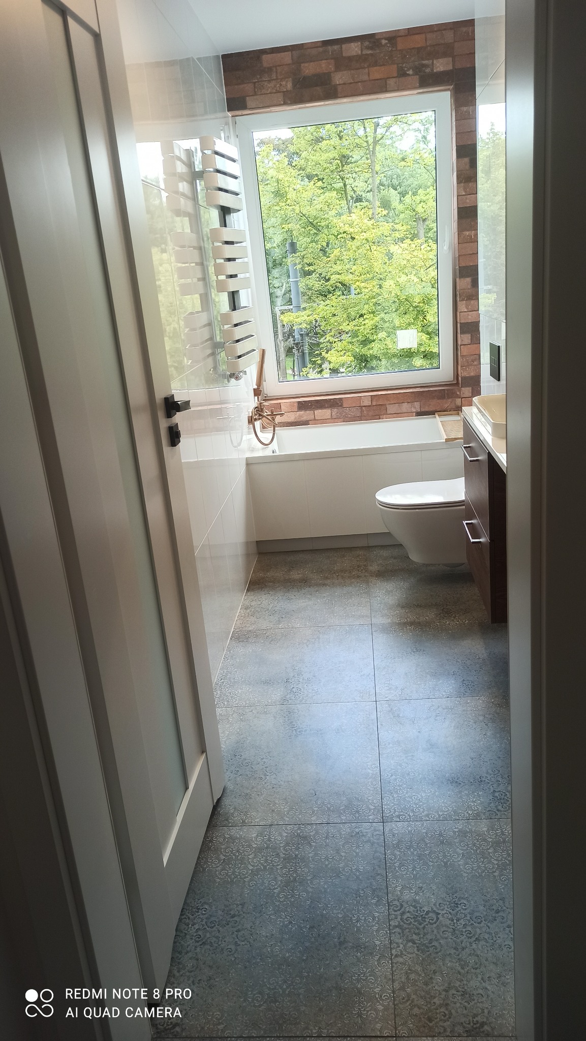 Nowoczesna łazienka z oknem wychodzącym na zieleń, szare płytki podłogowe z delikatnym wzorem, biała wanna obudowana płytkami, grzejnik drabinkowy i umywalka z szafką w kolorze ciemnego drewna.