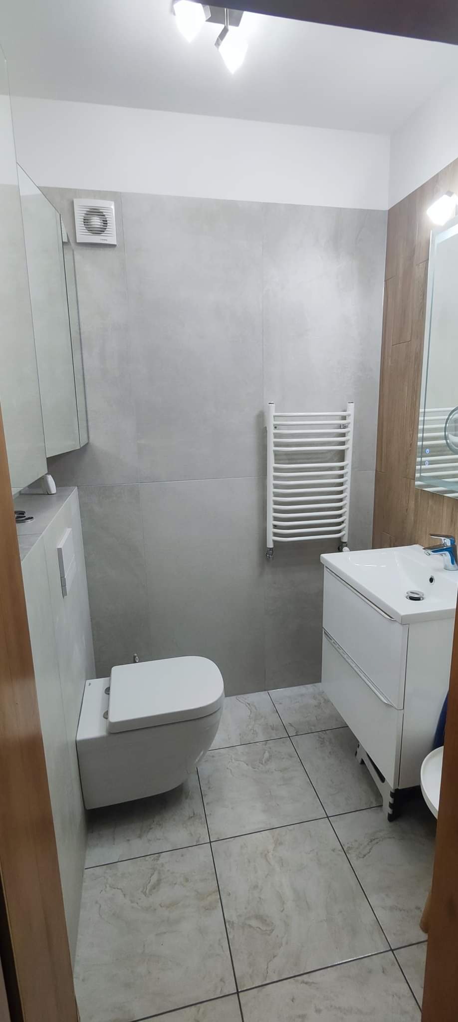 Wykończona łazienka w Gdyni z szarymi płytkami imitującymi marmur, wiszącą toaletą, grzejnikiem drabinkowym i białą szafką pod umywalką z chromowaną baterią.