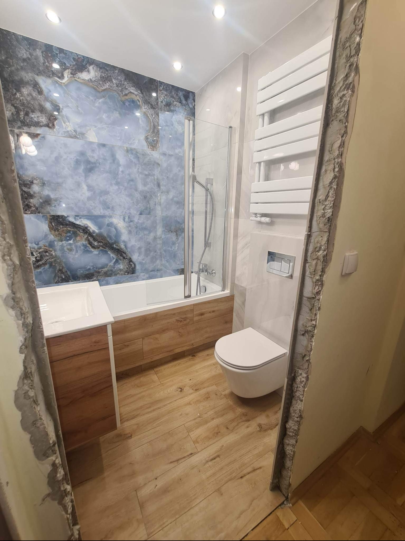 Nowoczesna łazienka w trakcie wykańczania: płytki imitujące marmur w odcieniach błękitu i szarości, drewnopodobna zabudowa wanny i umywalki, wisząca toaleta, grzejnik drabinkowy, widoczne surowe...