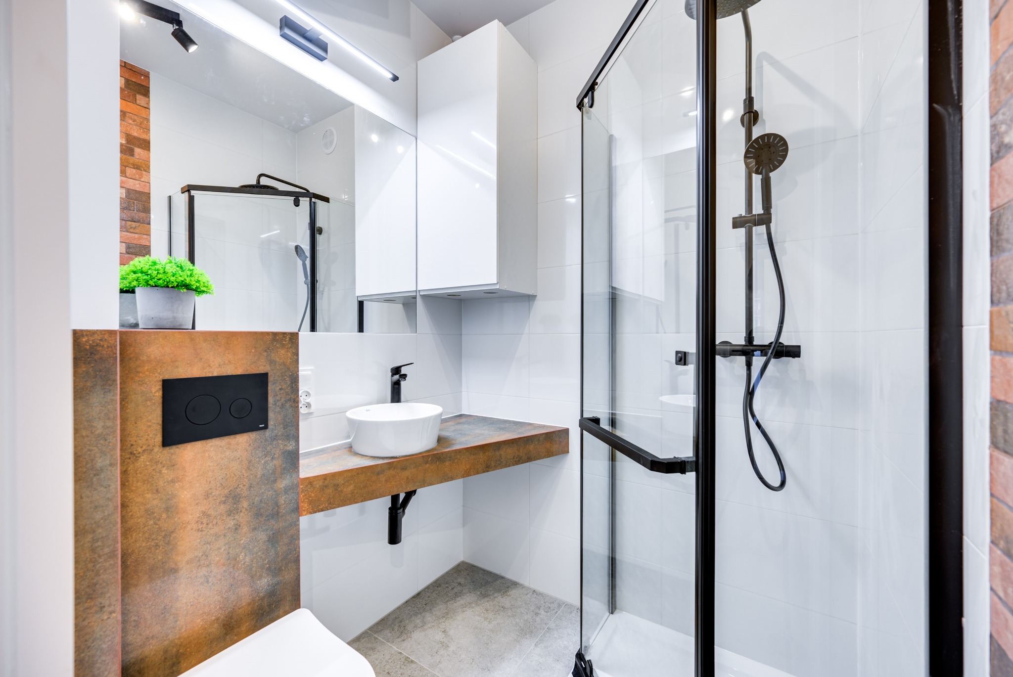 Nowoczesna łazienka z kabiną prysznicową z czarnymi ramami, umywalką nablatową na blacie imitującym rdzę, i białymi płytkami, akcentowana ceglaną ścianą i czarnym przyciskiem spłuczki.