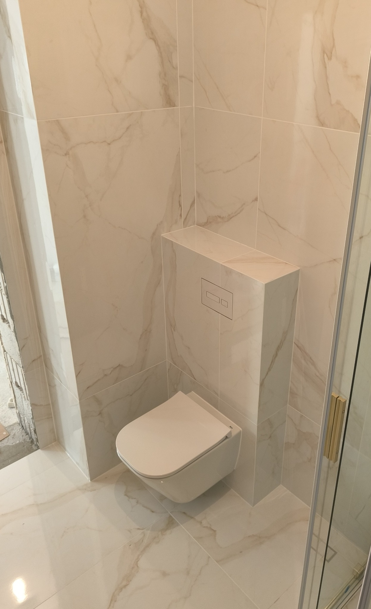 Wisząca toaleta z ukrytą spłuczką w zabudowie z płytek imitujących marmur, dopasowanych do ścian i podłogi w łazience po remoncie.