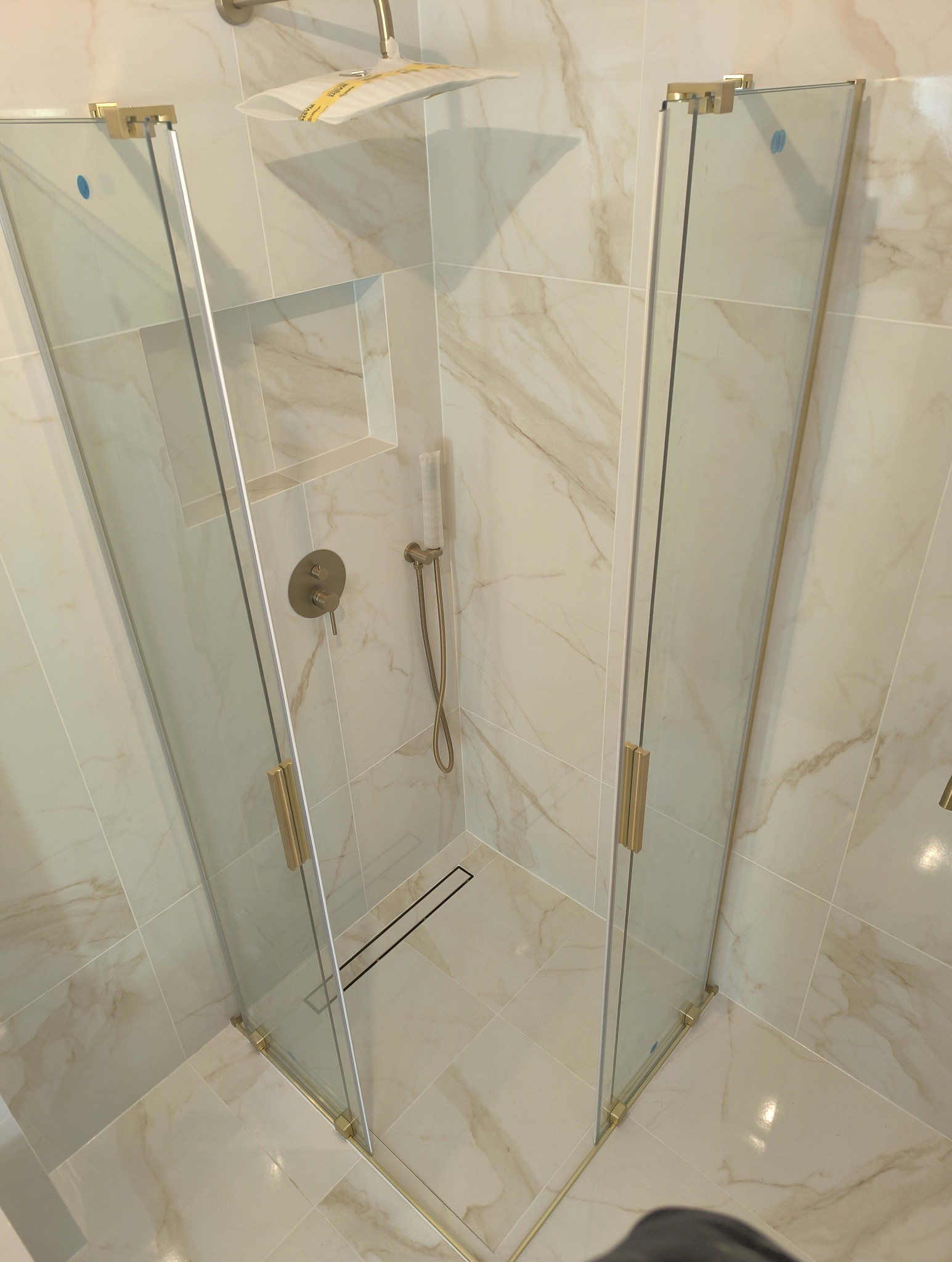 Kabina prysznicowa narożna, szklana z złotymi okuciami, zamontowana w wykończonej łazience z płytkami imitującymi marmur, widoczna bateria podtynkowa i deszczownica z folią ochronną oraz odpływ...