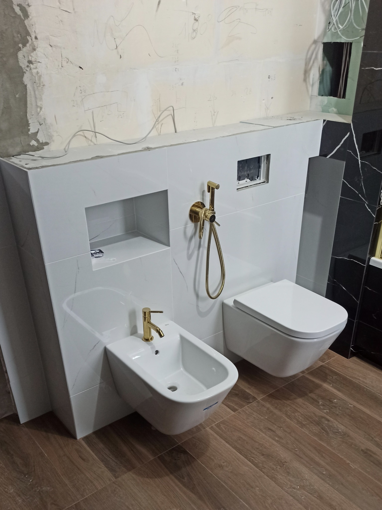 Bidet i toaleta podwieszana z armaturą w kolorze złota, zamontowane na białych płytkach imitujących marmur w łazience w Gdyni, ściana w tle w trakcie wykończenia.