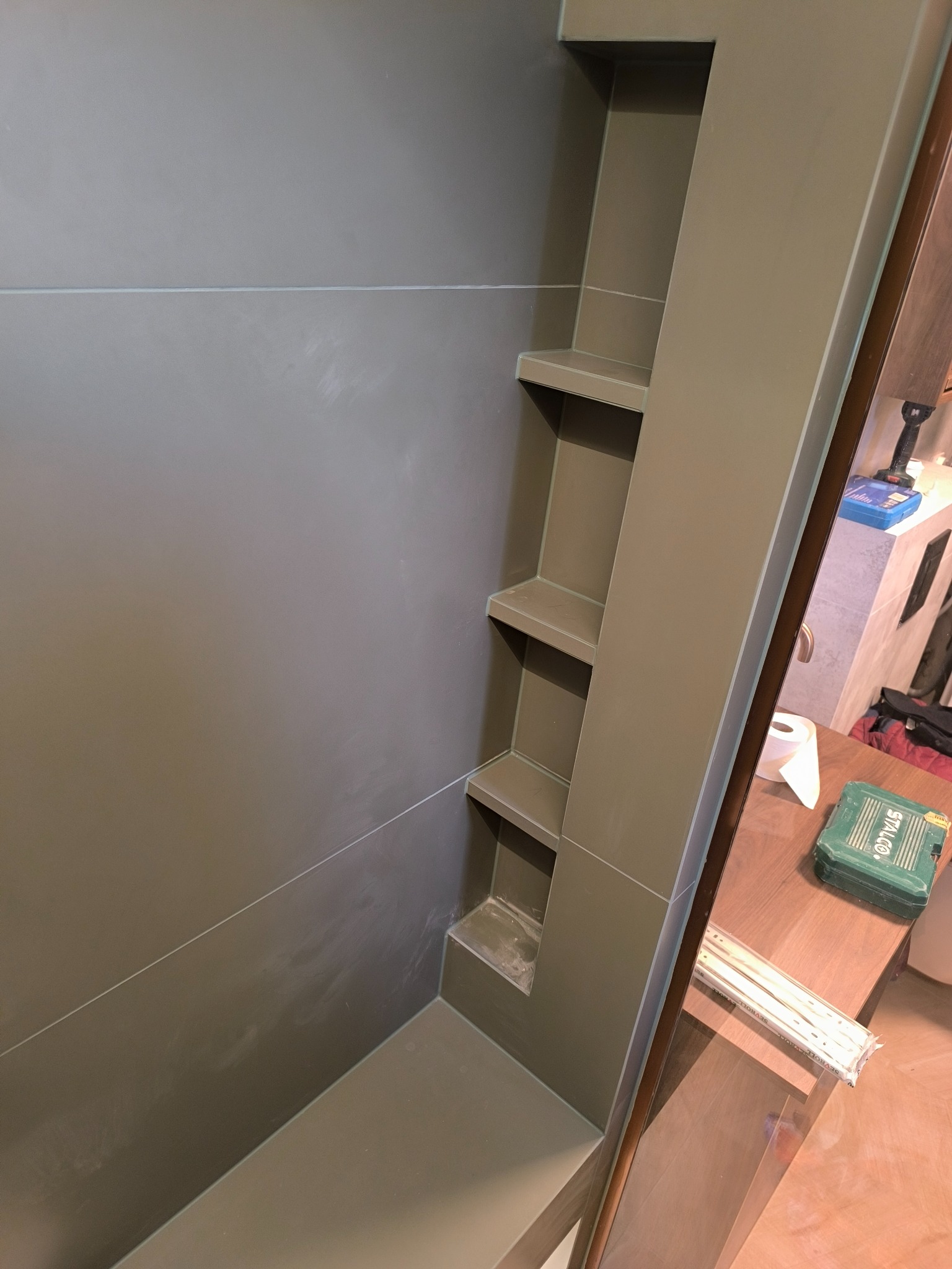 Wykończona wnęka z półkami w łazience, wyłożona płytkami w odcieniach szarości, z widocznymi fugami i narzędziami na blacie obok.