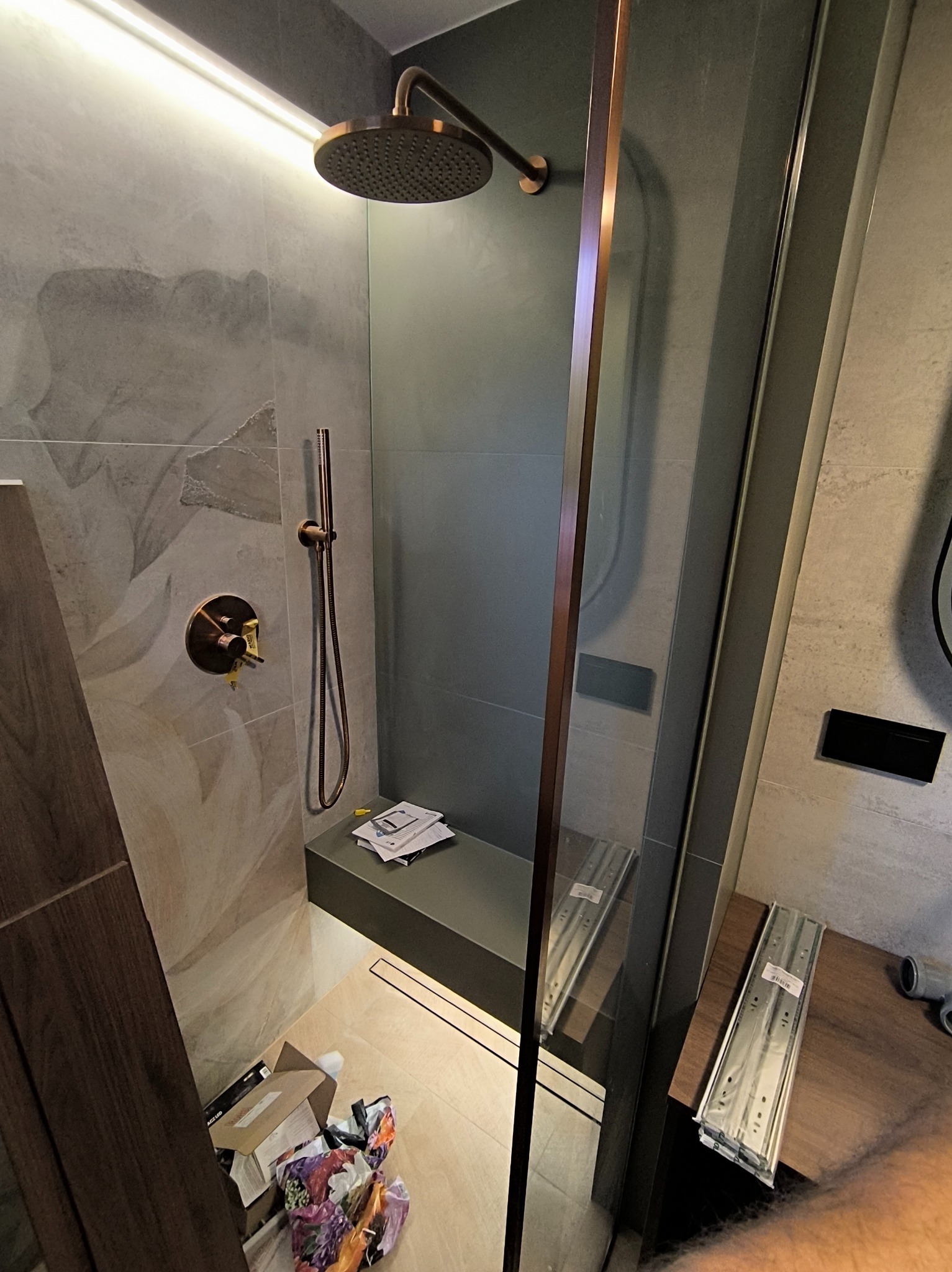 Nowoczesna kabina prysznicowa z deszczownicą w odcieniach szarości i miedzi. Widoczne elementy montażowe i akcesoria łazienkowe.