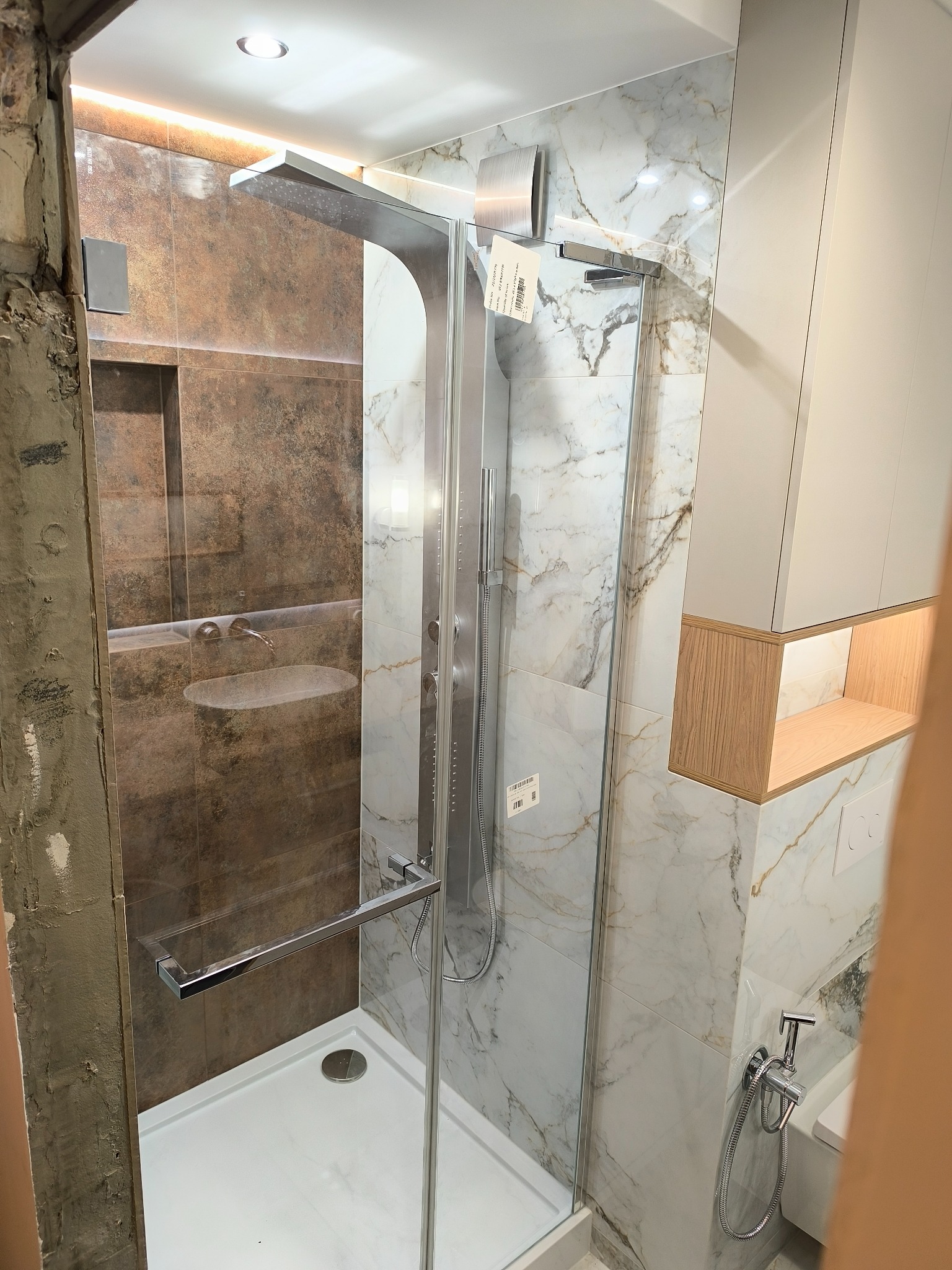 Wykończona kabina prysznicowa z deszczownicą, obłożona płytkami imitującymi marmur, z widoczną umywalką w tle i fragmentem ściany w stanie surowym.