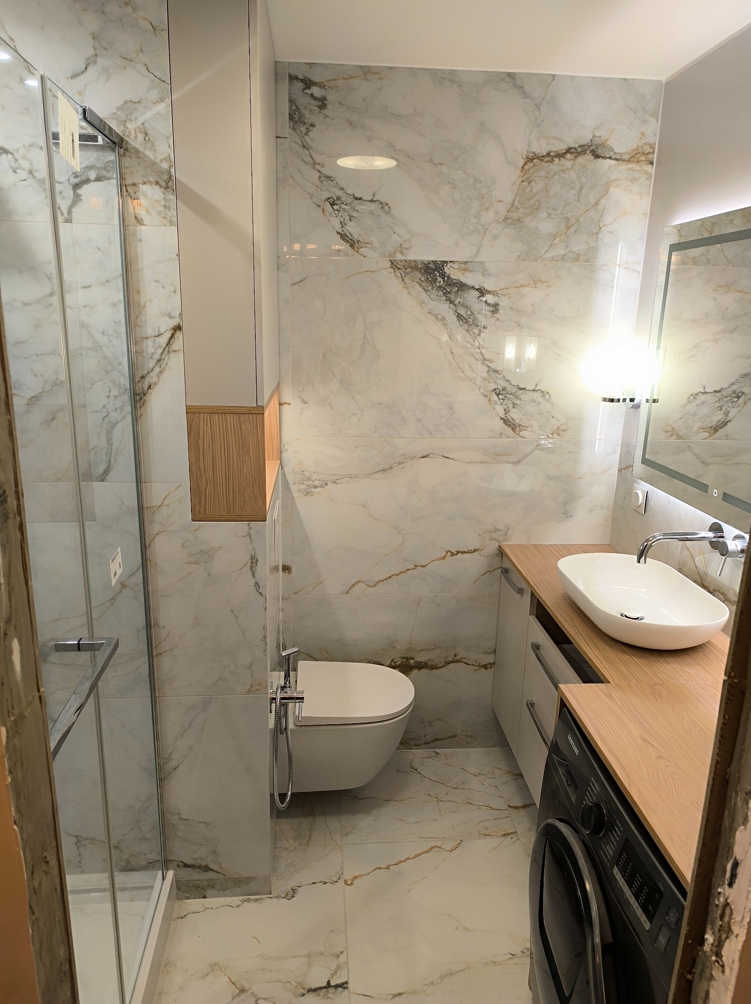 Elegancka łazienka z marmurowymi płytkami, kabiną prysznicową, umywalką nablatową i pralką. Jasne wnętrze z drewnianymi akcentami.