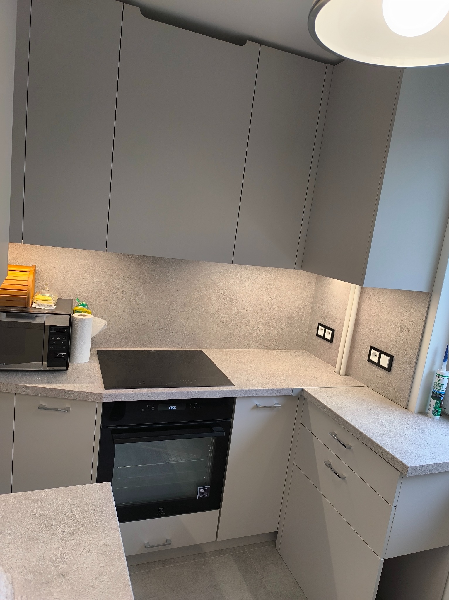 Jasna kuchnia z szarymi szafkami i blatem o teksturze kamienia. Widoczny piekarnik, płyta indukcyjna i gniazdka elektryczne w szarej ścianie nad blatem.