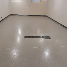 Jasnoszara, gładka podłoga z odpływem liniowym i listwami przypodłogowymi, widok na ścianę z ładowarką do samochodu elektrycznego.