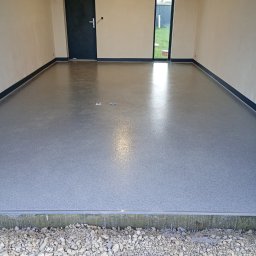 PAWEŁ RAKOWSKI STANDARD-BUD PLUS - Szara, gładka posadzka w surowym wnętrzu z drzwiami i oknem. Widoczny fragment podłoża z kamieniami. Minimalistyczny design, surowy stan wykończenia.