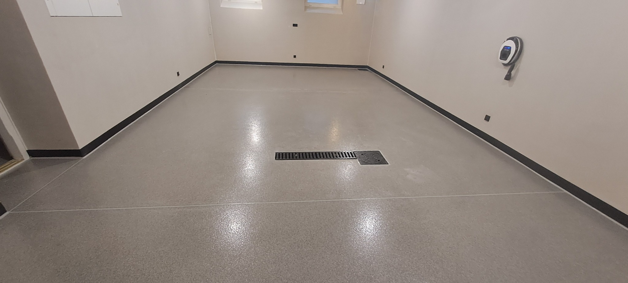 Jasnoszara, gładka podłoga z odpływem liniowym i listwami przypodłogowymi, widok na ścianę z ładowarką do samochodu elektrycznego.