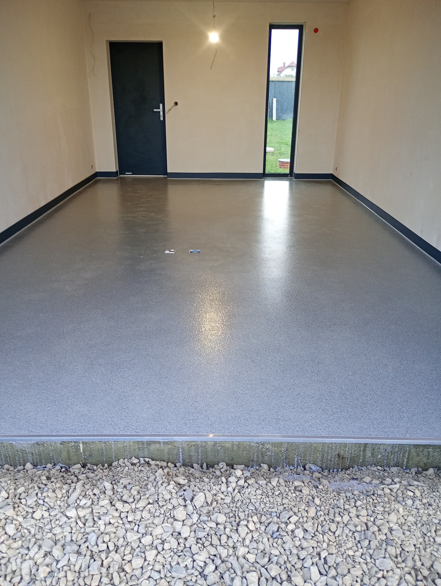 Szara, gładka posadzka w surowym wnętrzu z drzwiami i oknem. Widoczny fragment podłoża z kamieniami. Minimalistyczny design, surowy stan wykończenia.