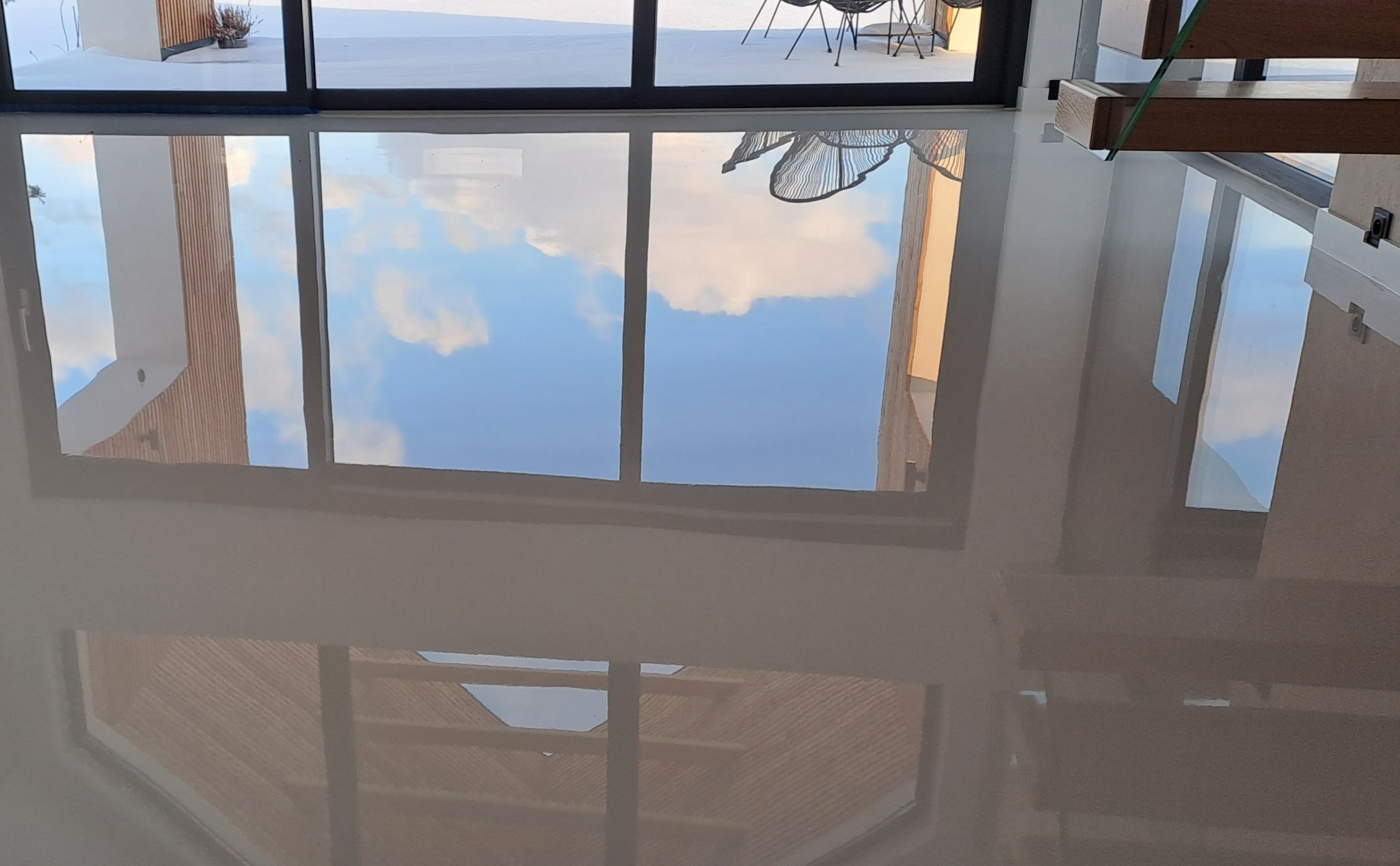 Błyszcząca, beżowa podłoga z żywicy epoksydowej odbija okno z widokiem na błękitne niebo i chmury. Widoczne fragmenty schodów i mebli.