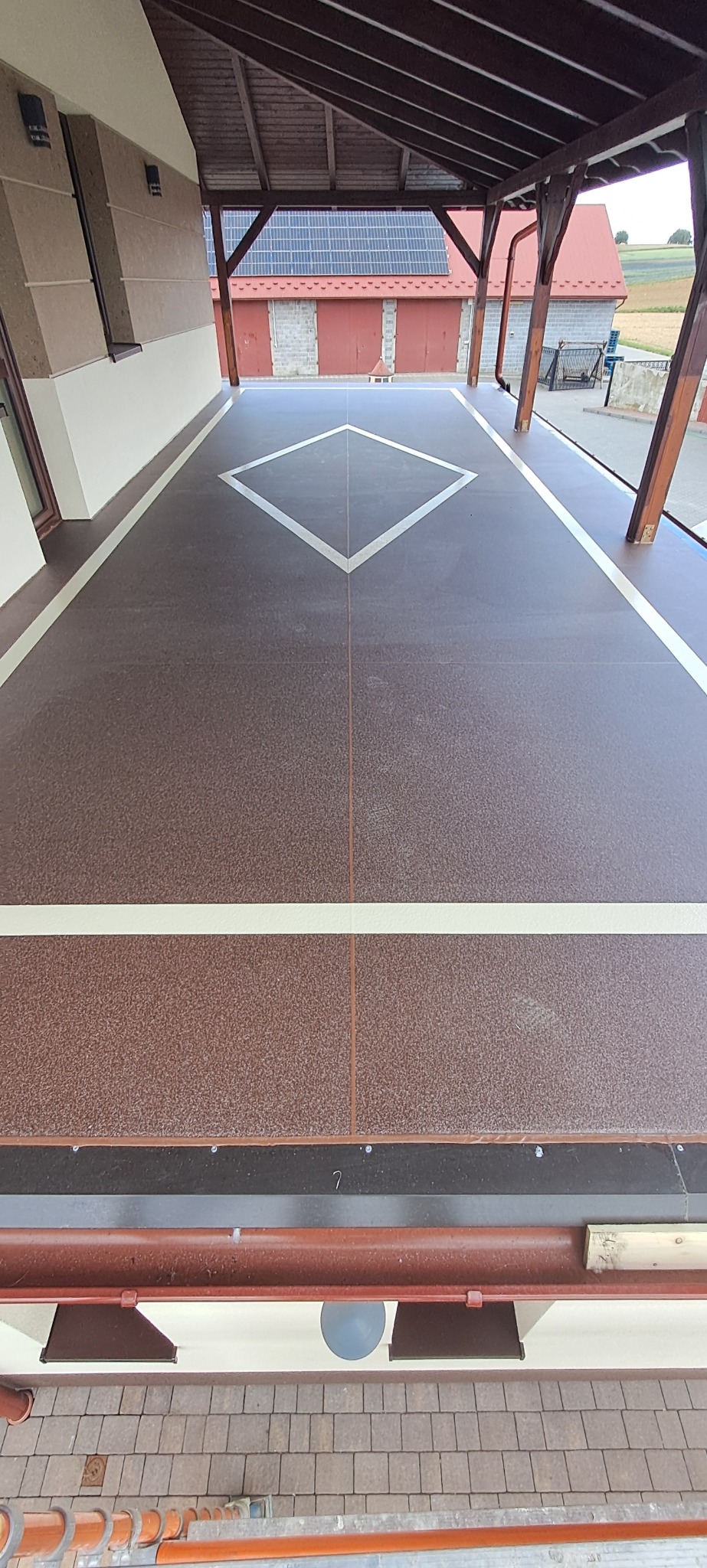 Balkon z brązową posadzką żywiczną, zdobioną białym rombem i liniami. Zadaszenie z drewnianą konstrukcją. Widok z góry na wykończoną powierzchnię.