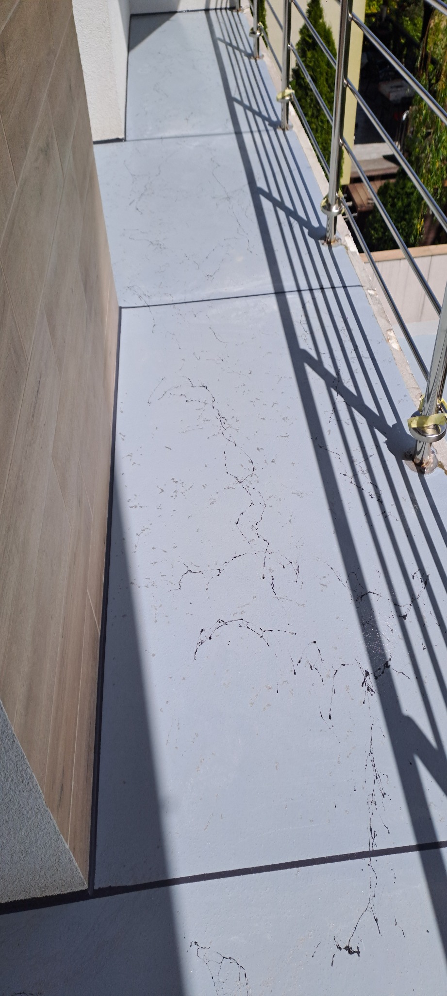 Szara posadzka żywiczna na balkonie z metalową balustradą, widoczne cienie balustrady i wzór imitujący marmur. Fragment ściany wykończonej płytkami drewnopodobnymi.