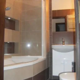 Ukończona łazienka z beżowo-brązowymi płytkami, narożną wanną z parawanem, białą umywalką z szafką i wiszącą toaletą.