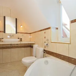 Jasna łazienka na poddaszu z beżowymi płytkami, umywalką nablatową, toaletą i wanną narożną, oświetlona dwoma kinkietami i światłem dziennym z okna dachowego.