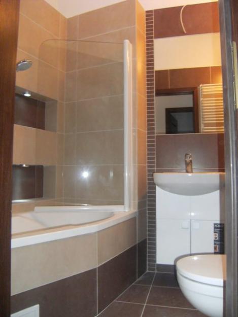 Ukończona łazienka z beżowo-brązowymi płytkami, narożną wanną z parawanem, białą umywalką z szafką i wiszącą toaletą.
