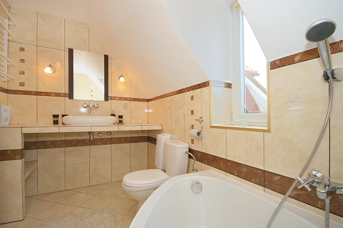 Jasna łazienka na poddaszu z beżowymi płytkami, umywalką nablatową, toaletą i wanną narożną, oświetlona dwoma kinkietami i światłem dziennym z okna dachowego.