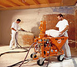 Dwóch pracowników w białych koszulkach aplikuje tynk maszynowy na ścianę za pomocą pomarańczowej maszyny do tynkowania. Jeden pracownik trzyma wąż i nakłada tynk, drugi dosypuje materiał z worka...
