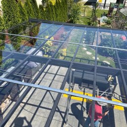 PIR-TECH - Montaż szklanego zadaszenia nad tarasem z widokiem na ogród, pracownik w czerwonej koszulce mocuje elementy konstrukcji.