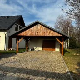 Drewniana wiata garażowa z ciemnym dachem przy domu z jasną elewacją, obok czerwony samochód. Podłoże wyłożone drobnym kamieniem. W tle las.