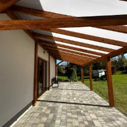 Zadaszony taras z drewnianą konstrukcją i jasną markizą, wyłożony kostką brukową. Widok na ogród i błękitne niebo w tle. Eleganckie połączenie drewna i nowoczesnego designu.