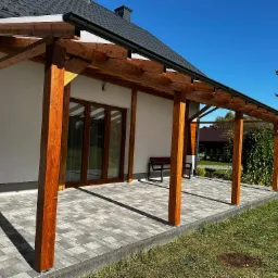 Drewniana pergola zadaszona, z widocznymi elementami konstrukcyjnymi i fragmentem dachu budynku w Terliczce. Na pierwszym planie szara kostka brukowa.