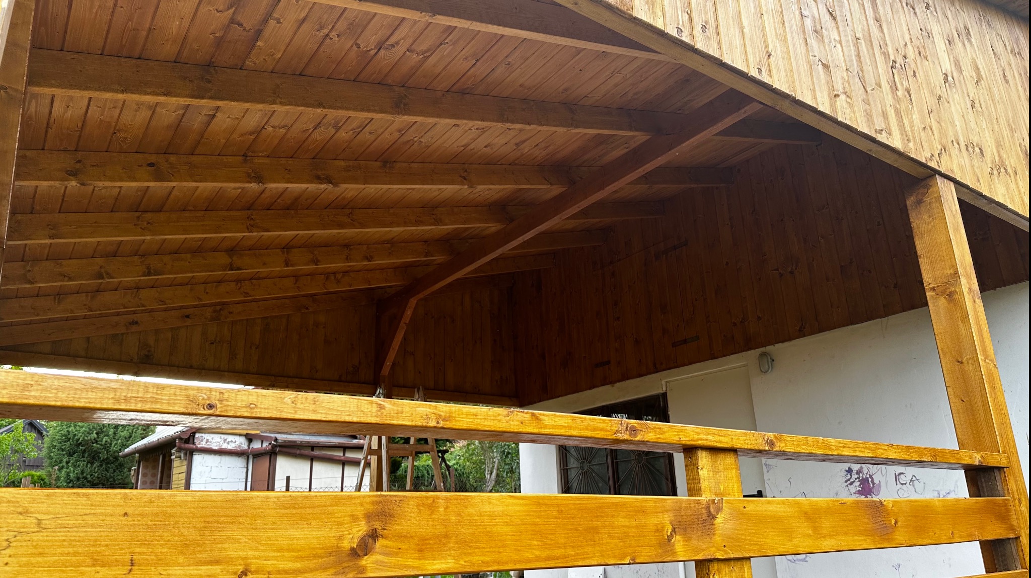 Zadaszenie drewniane z widoczną strukturą desek, świeżo pomalowane na złocisty kolor, z elementami konstrukcyjnymi i balustradą, na tle budynku z białymi ścianami i zielenią w oddali.