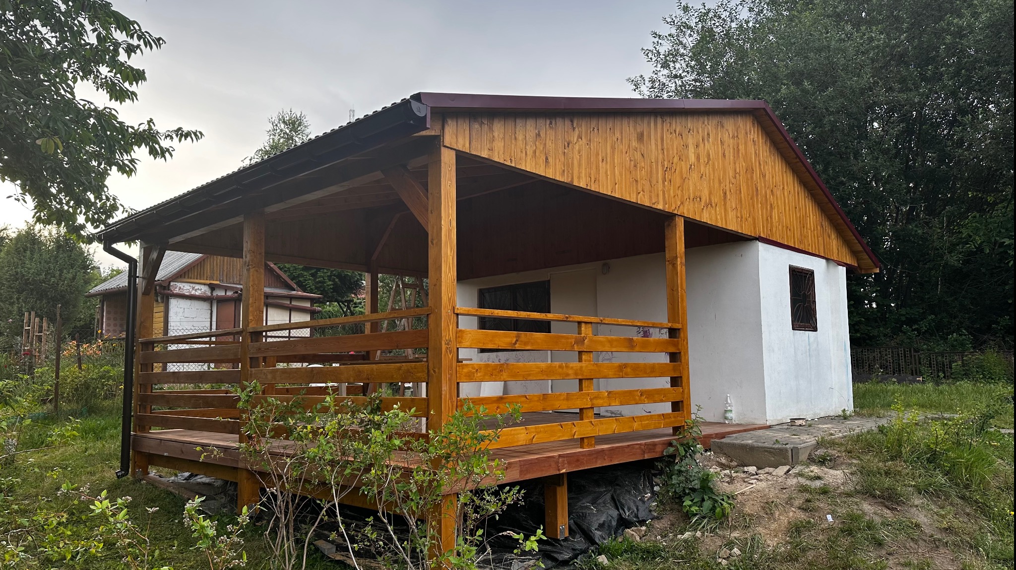 Drewniana konstrukcja z zadaszeniem, połączona z budynkiem, z widoczną drewnianą podłogą i balustradą, otoczona zielenią.