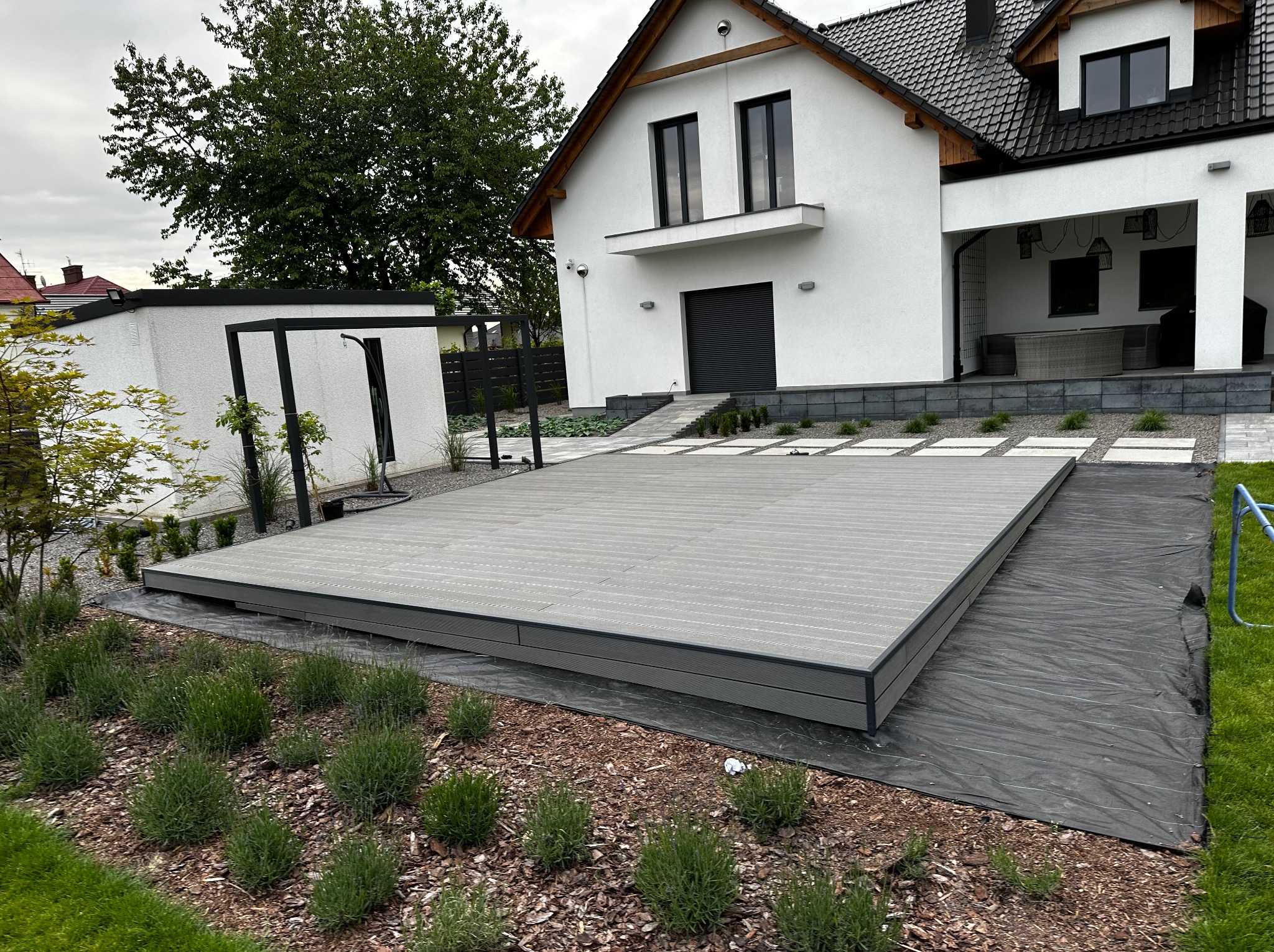 Nowoczesny taras kompozytowy w kolorze szarym, na przygotowanym podłożu z agrowłókniny, z widocznymi elementami ogrodu i fragmentem białego domu w tle.