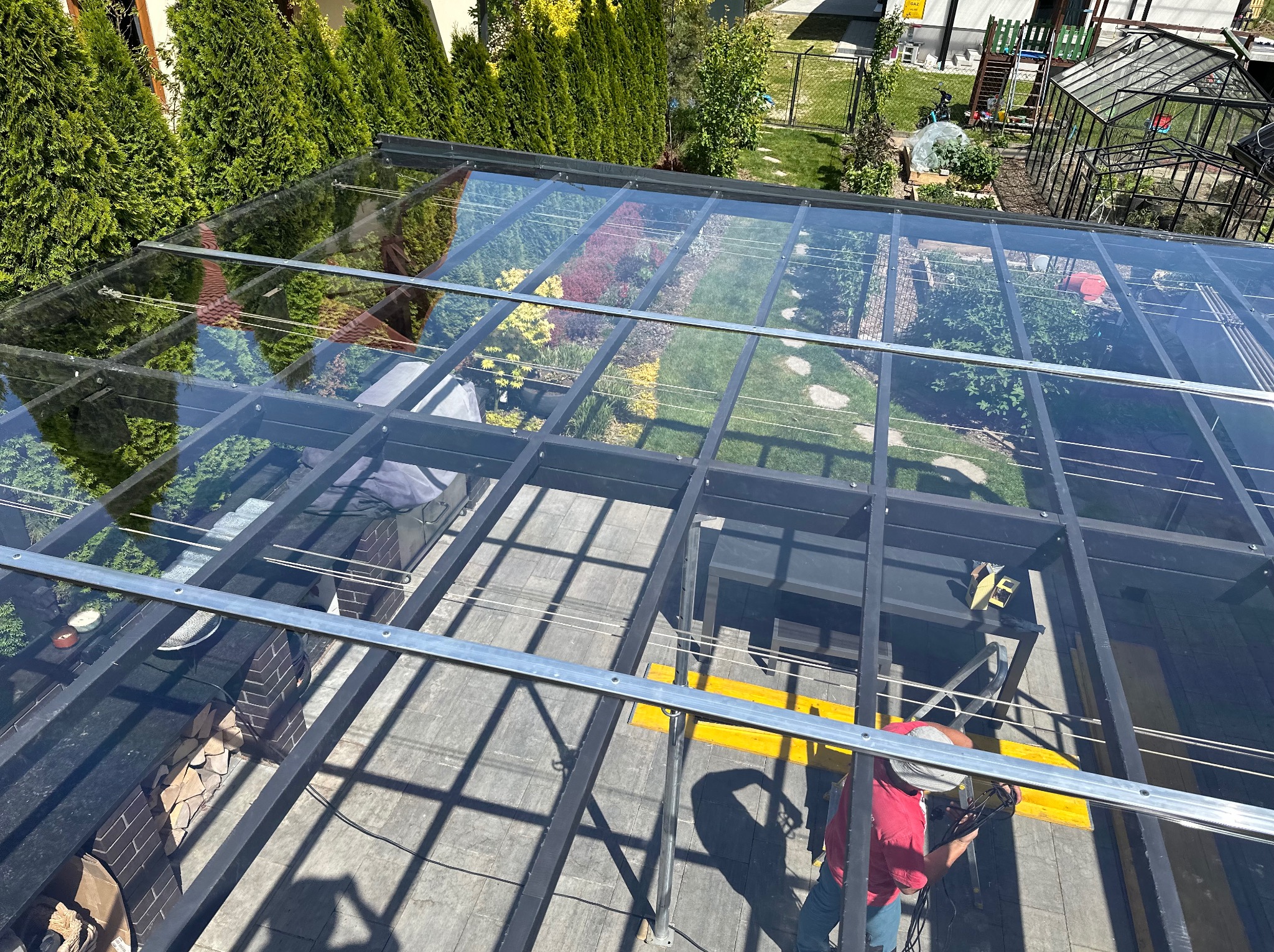 Montaż szklanego zadaszenia nad tarasem z widokiem na ogród, pracownik w czerwonej koszulce mocuje elementy konstrukcji.