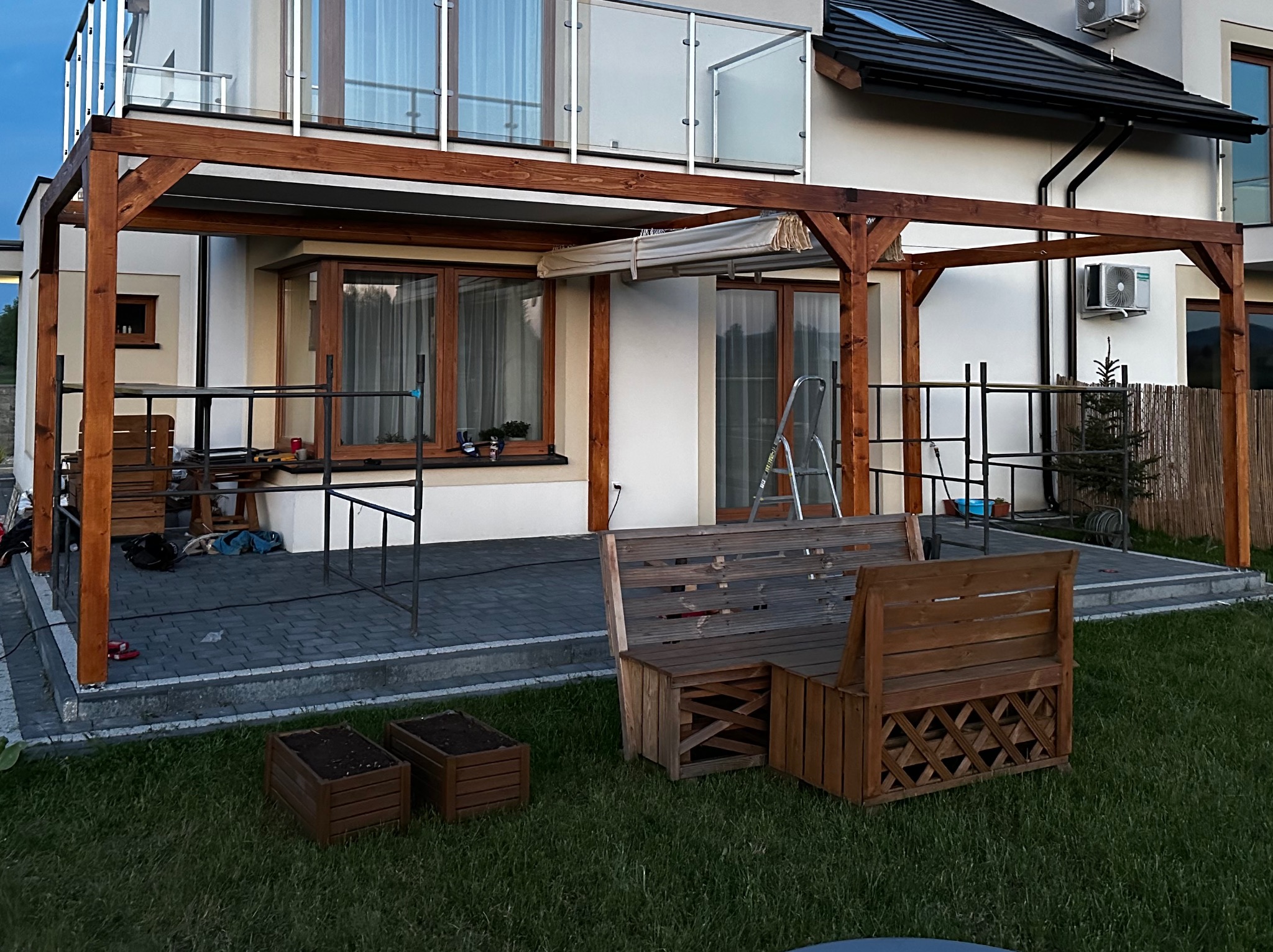 Drewniana pergola w Terliczce, częściowo zacieniona roletą, z widocznymi meblami ogrodowymi z palet i donicami na trawniku, na tle nowoczesnego domu z balkonem.
