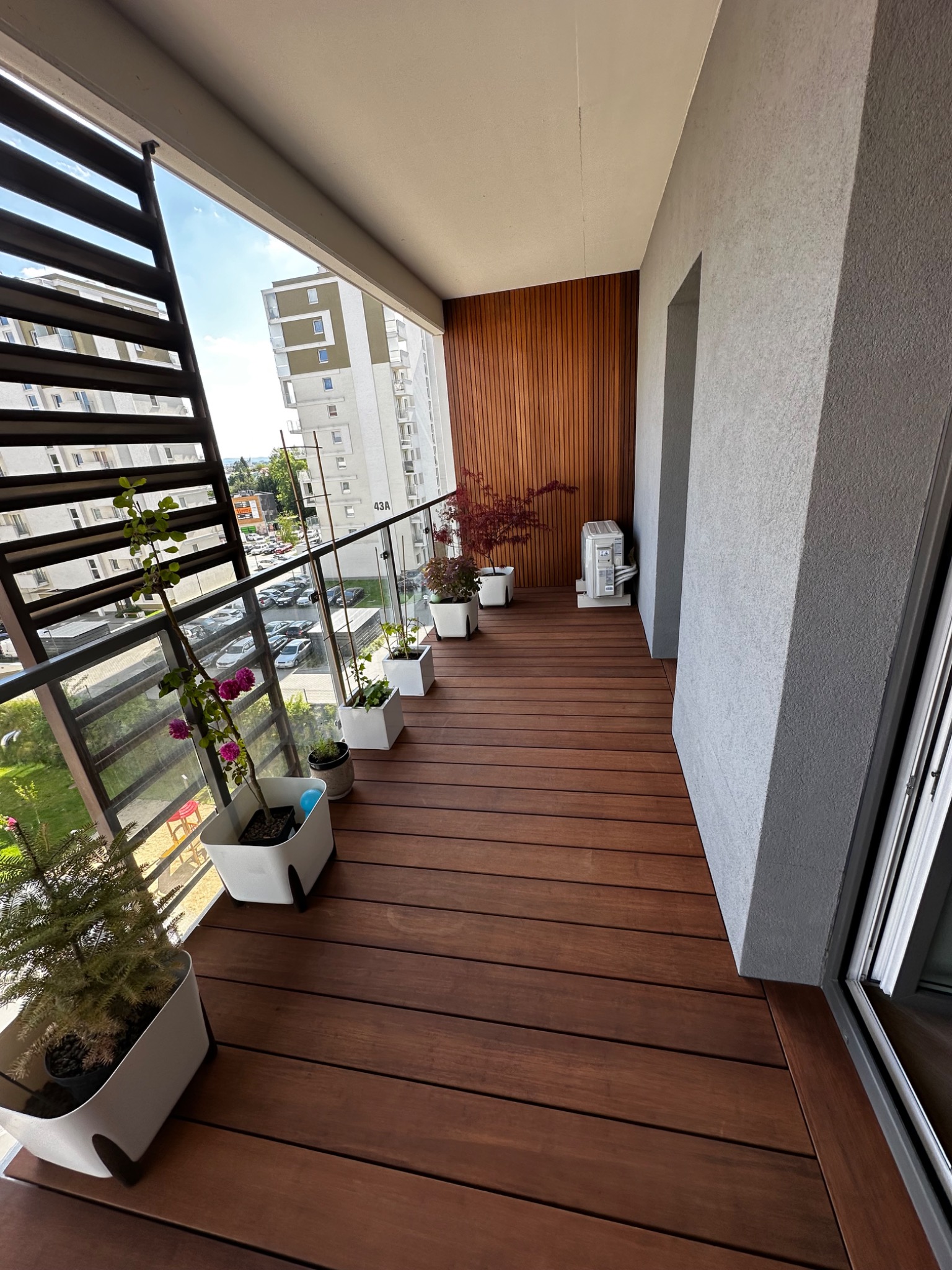 Długi balkon z brązową podłogą kompozytową, szklaną balustradą i donicami z roślinami. Na końcu balkonu widoczna drewniana ściana i klimatyzator.