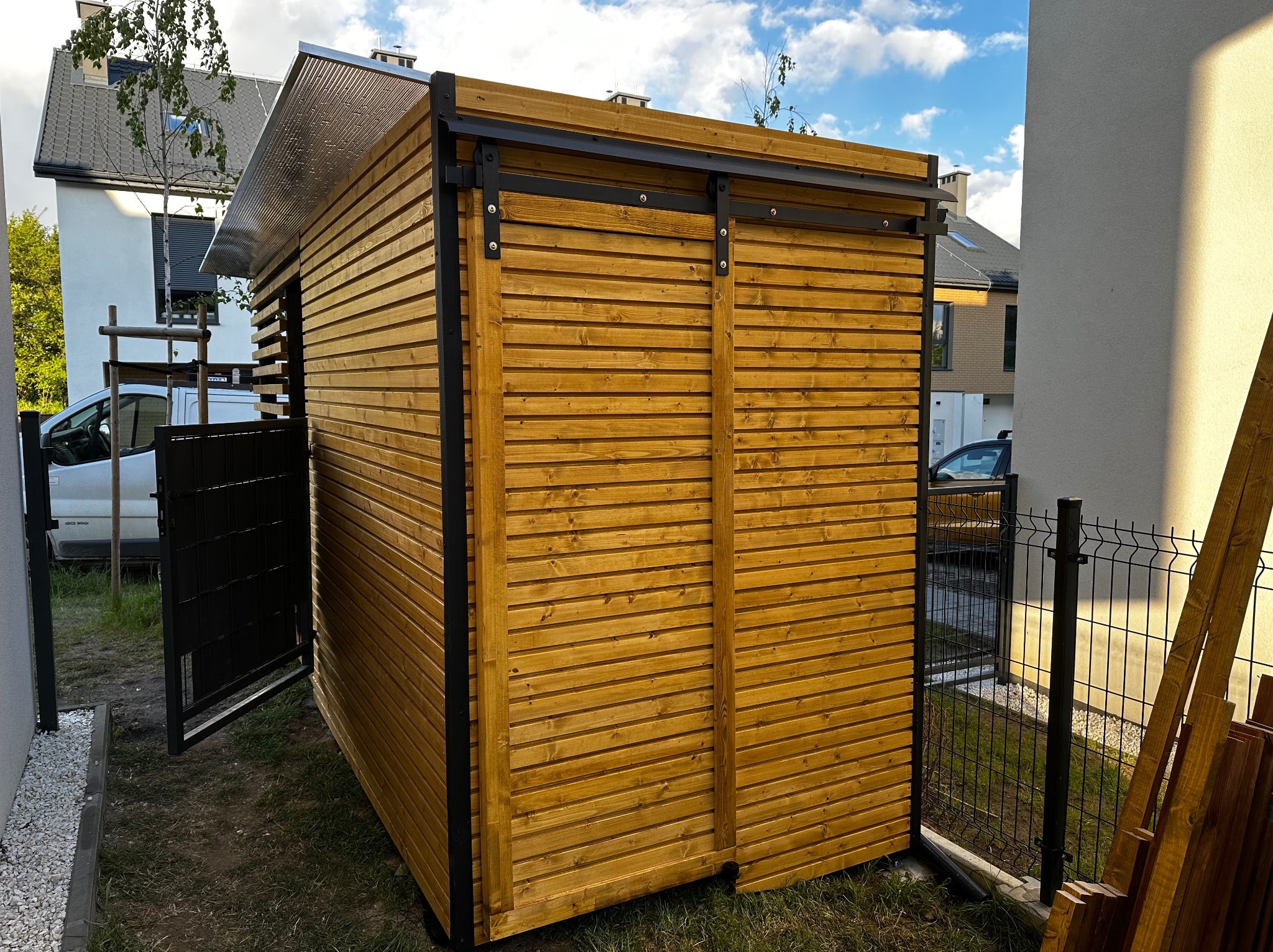 Drewniana altana z przesuwnymi drzwiami, zadaszona blachą, na tle zielonego trawnika i zabudowy jednorodzinnej. Otwarta brama w tle. Elementy metalowe w kolorze czarnym.