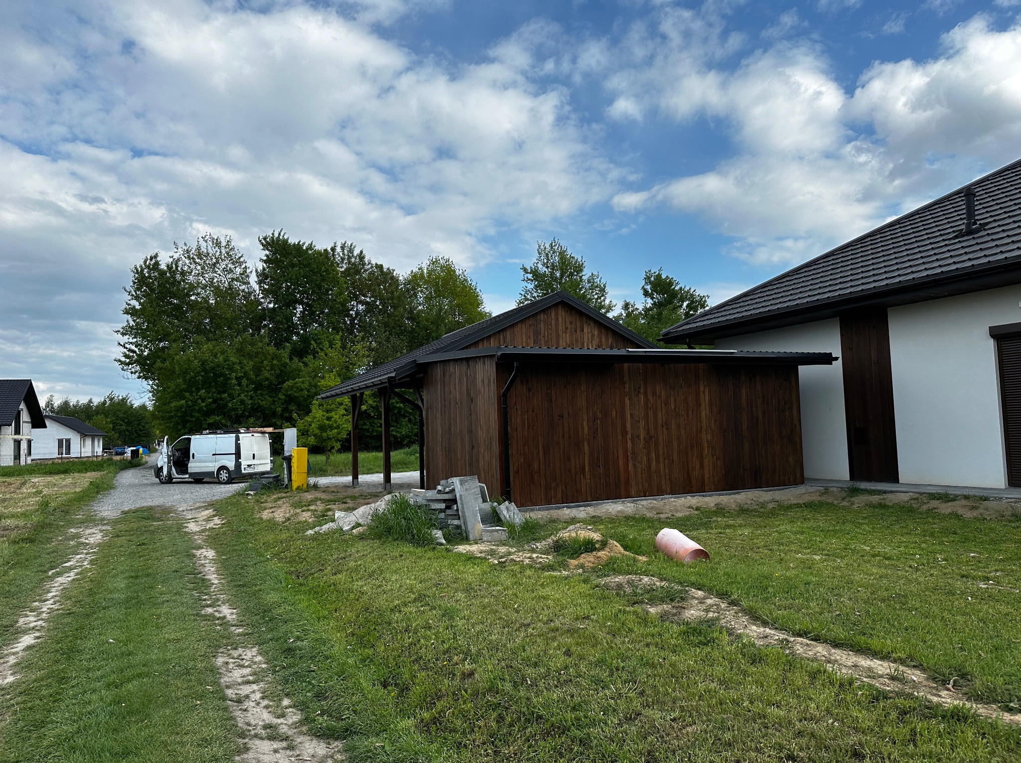 Drewniana pergola zadaszona blachą, widoczna z boku, na tle zieleni i błękitnego nieba z chmurami. Biały bus w oddali. Częściowo widoczny budynek z białymi ścianami.