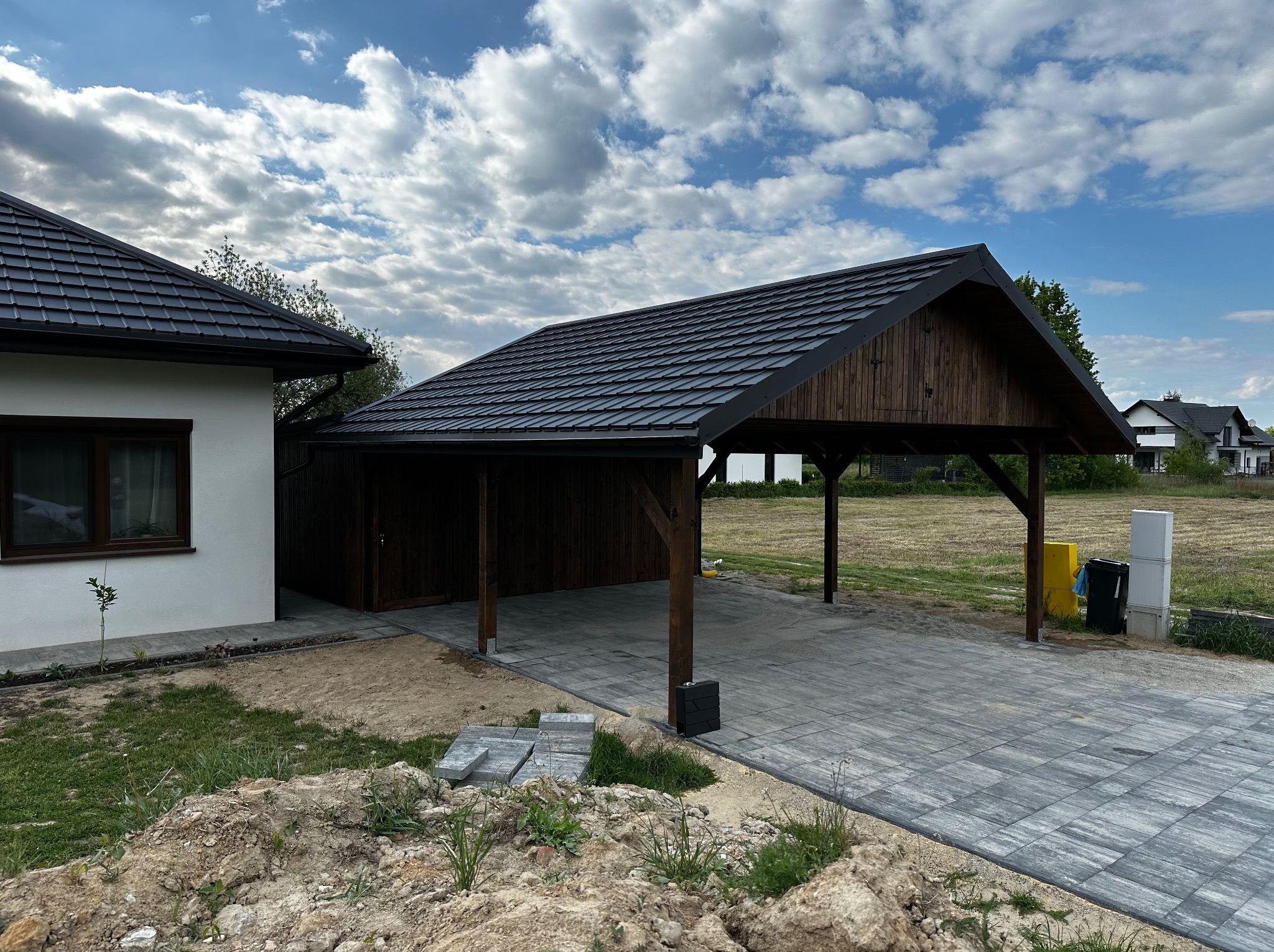 Drewniana wiata garażowa z ciemną dachówką, połączona z budynkiem. Wybrukowany podjazd z szarej kostki. Elementy drewniane w odcieniu brązu, nowoczesna architektura.