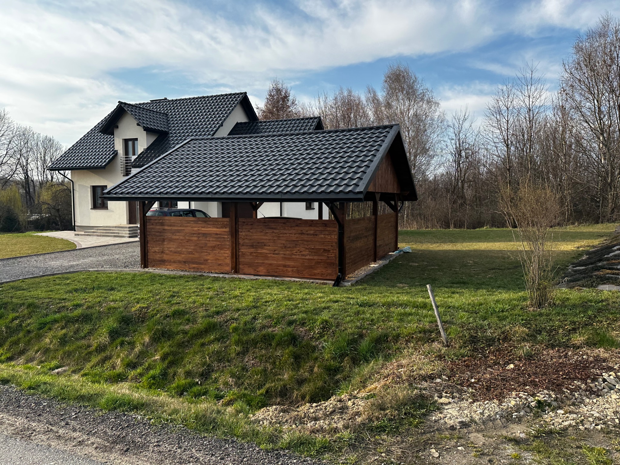 Drewniana pergola z czarnym dachem przylega do jasnego domu z ciemnym dachem. Widoczne drzewa w tle, zielony trawnik i szara droga.