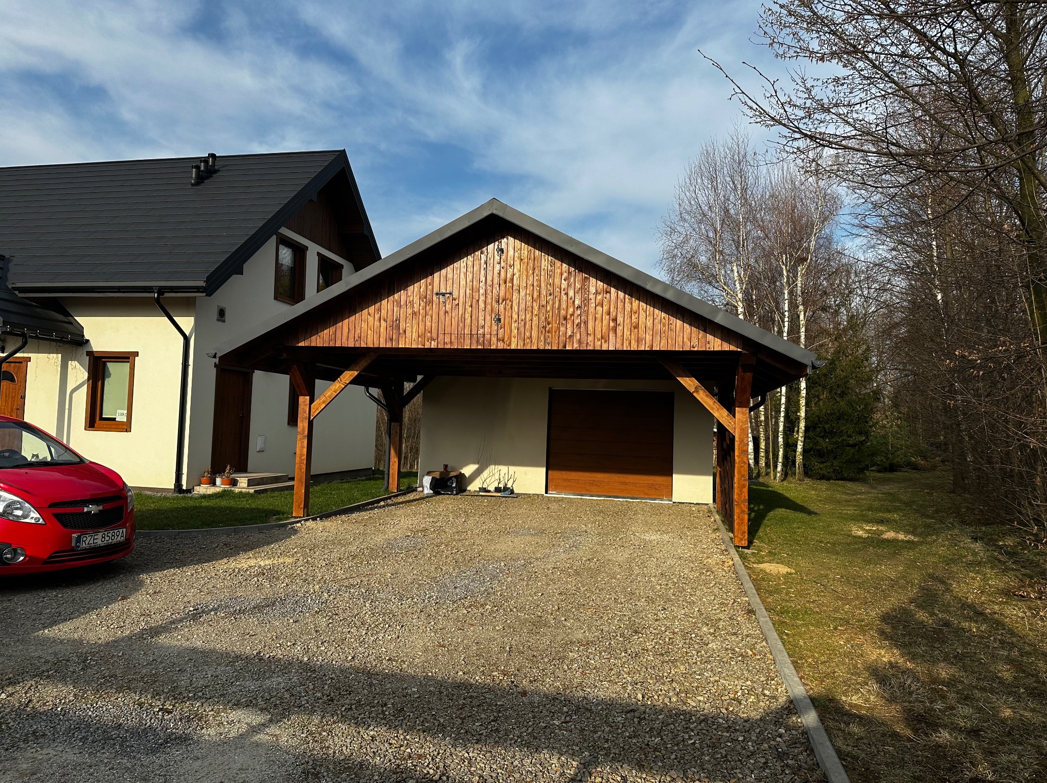 Drewniana wiata garażowa z ciemnym dachem przy domu z jasną elewacją, obok czerwony samochód. Podłoże wyłożone drobnym kamieniem. W tle las.