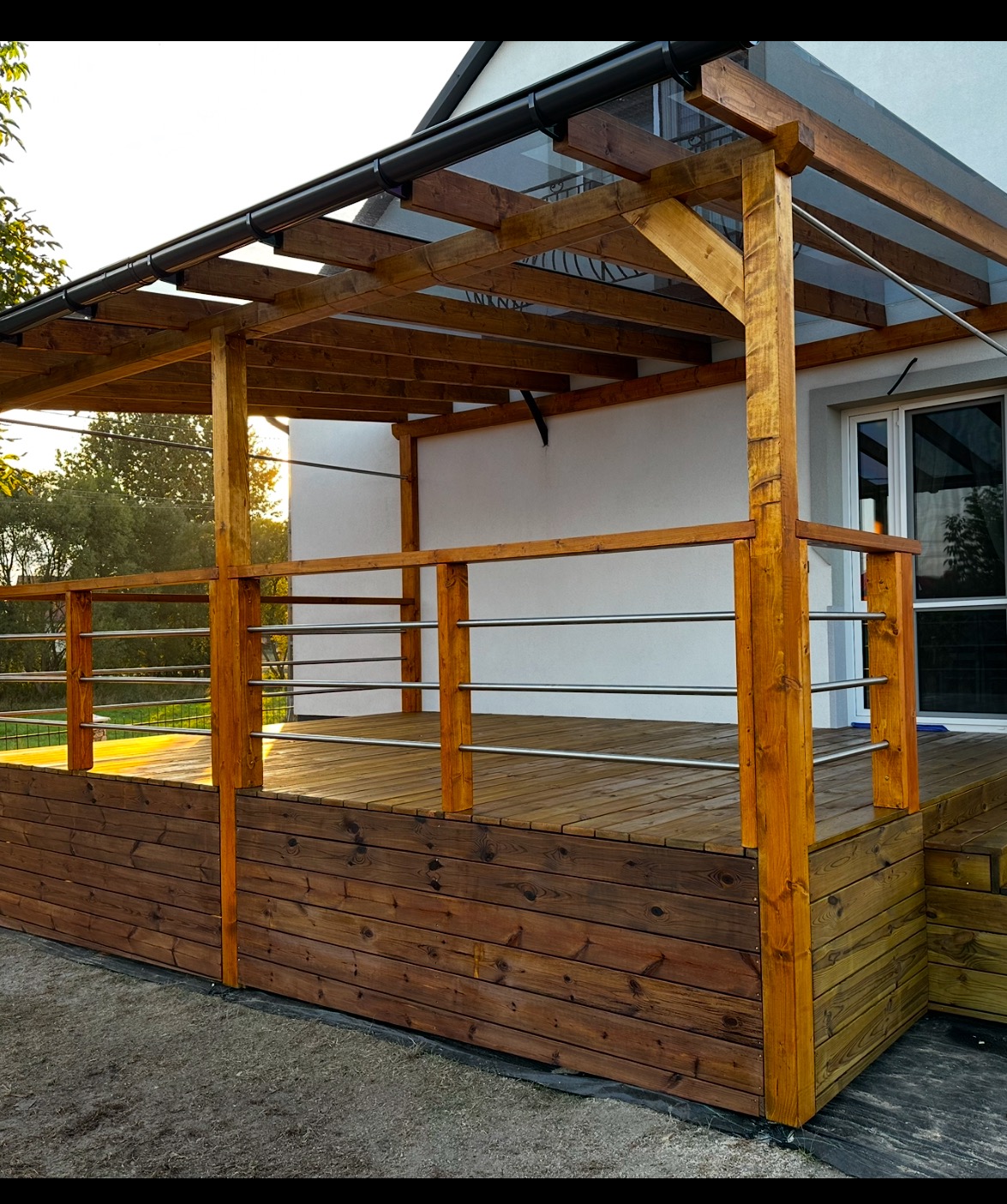 Drewniany taras z zadaszeniem z przezroczystego tworzywa, otoczony niską balustradą z metalowymi prętami. Widoczny fragment elewacji domu z oknem.